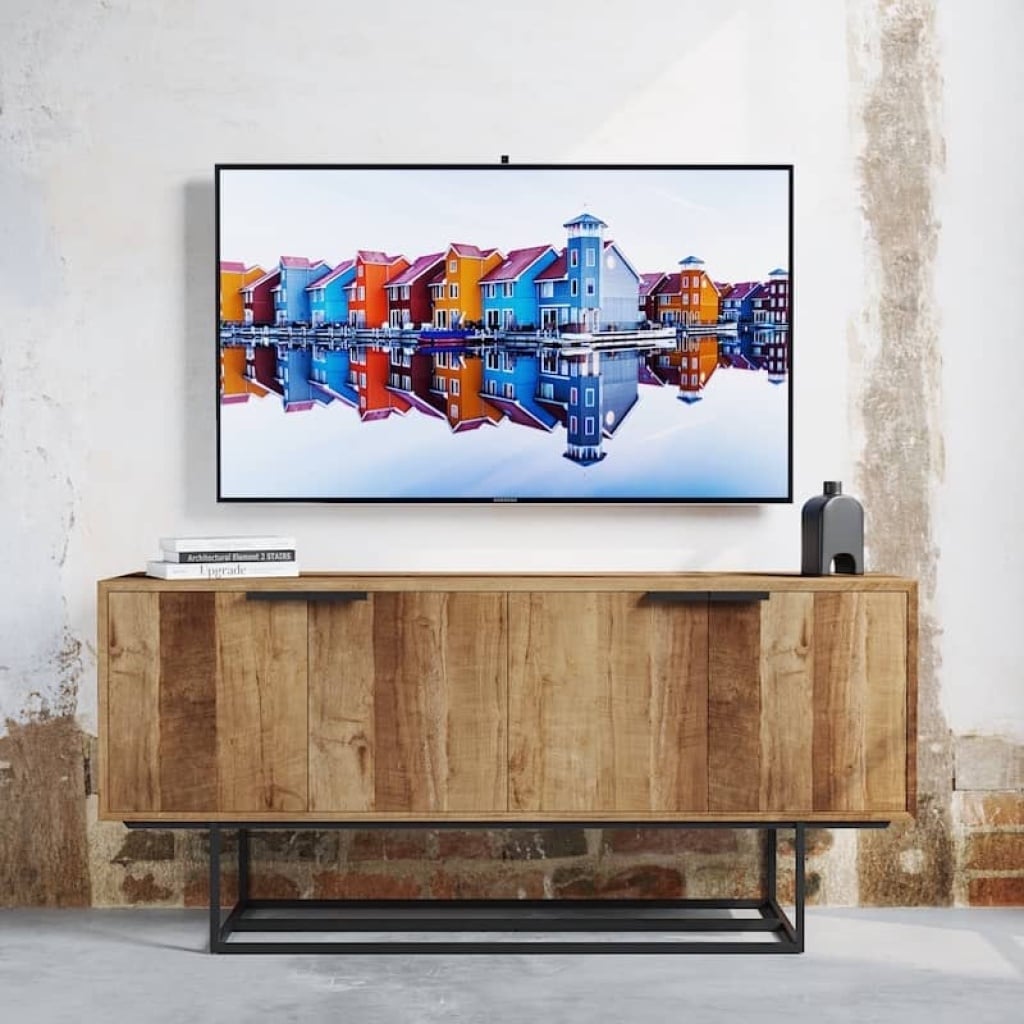 A wall-mounted TV displays a photo of colorful houses reflected on water. Below, the Parisian Factory 4 Door 72″ TV Console, designed with a wooden finish and metal frame, holds books and a small decor item. The background wall has a weathered look.