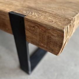 Close-up view of the Hudson Coffee Table, featuring a wooden top with a rough, unfinished edge and a black metal leg, set on a concrete floor.