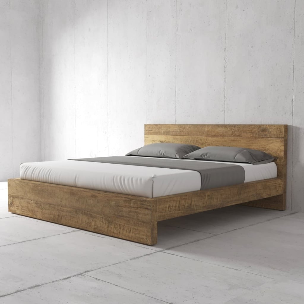 A minimalist bedroom features a wooden bed frame with a headboard, a white mattress, and grey pillows. The room has bare concrete walls and floor.
