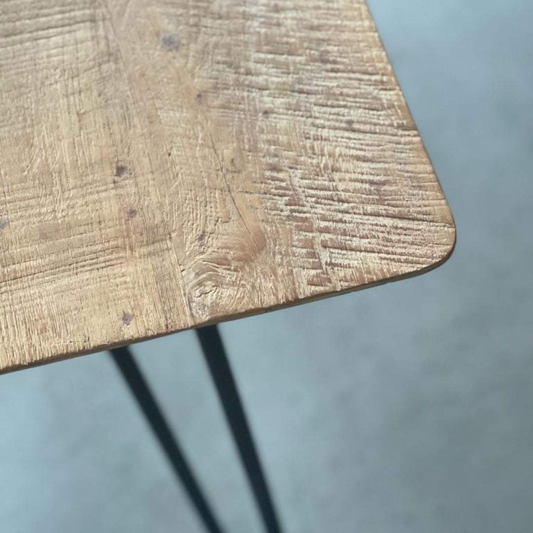 63 CABRILLO TABLE NATURAL (IMAGE 2) Close-up of the Cabrillo 63" Dining Table corner with a textured wooden surface and metal legs, viewed from above against a blurred grey background.