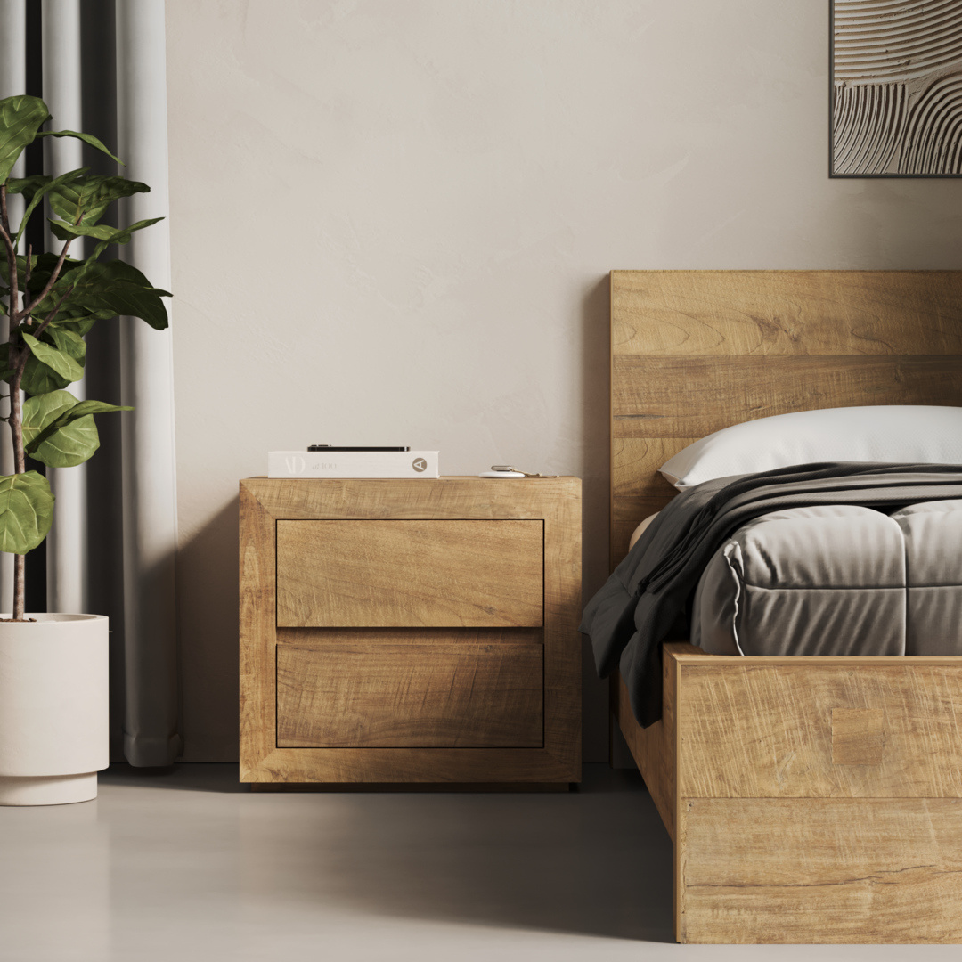 A wooden bed with white and gray bedding is next to a matching Lugano Nightstand adorned with books and a small decor piece. A potted plant is on the left side, and part of a wall art piece is visible on the right.