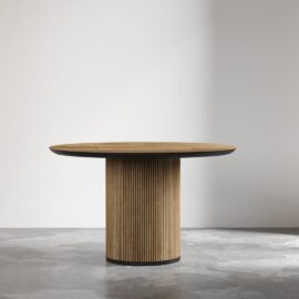 The Bolivar Round Dining Table with a cylindrical base stands in an empty room with a light-colored wall and a concrete floor.
