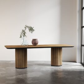 The Bolivar Dining Table, featuring two cylindrical bases, stands in a minimalist room, adorned with a glass vase holding branches and a ceramic pot.