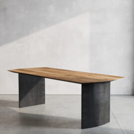 The Atelier Dining Table, featuring a modern rectangular wooden top and two black metal legs, stands on a light-colored floor against a plain wall, illuminated by sunlight.