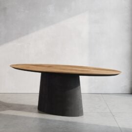 The Atelier Oval Dining Table, featuring a modern wooden design and a thick black cylindrical base, is positioned on a light gray concrete floor and set against a plain light gray wall.