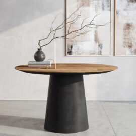 The Atelier Round Dining Table with a black base graces a minimalist room. Adorning the table are a vase filled with branches, several books, and a candle holder. On the wall behind hang two abstract paintings.