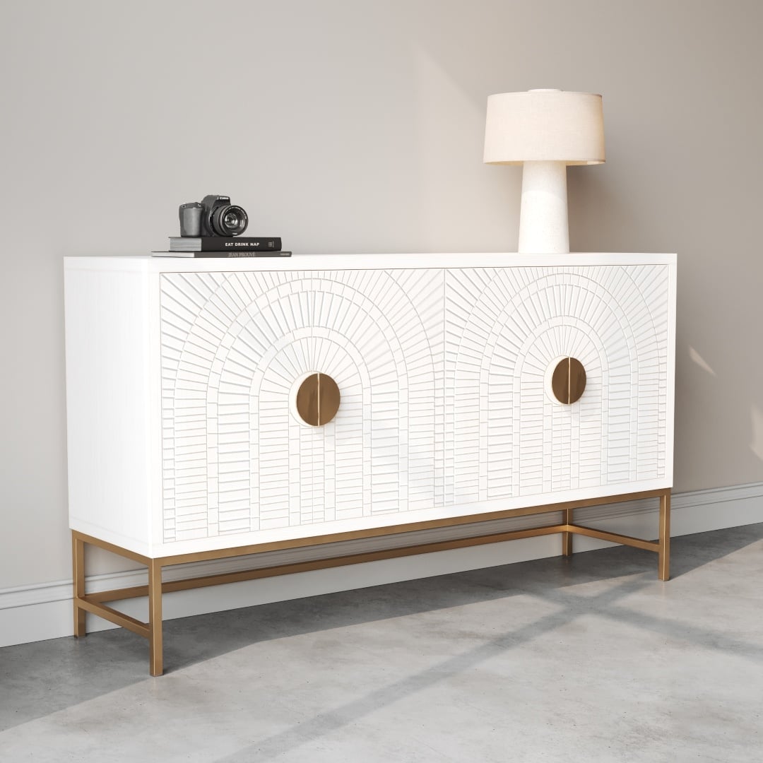 Coral 60 Side Cabinet -c The Coral 60" Sideboard features a white finish with an arched pattern on its doors, complemented by round golden handles and a golden base. It sits against a light-colored wall on a concrete floor, with a lamp and camera elegantly placed on top.