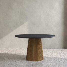 The Osaka Round Dining Table, featuring a dark top and a textured, cylindrical wooden base, stands on a speckled floor against a plain, light-colored wall.