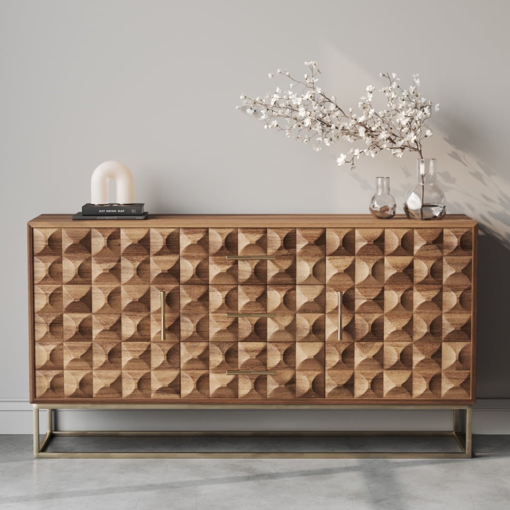 A Wadi 58" Sideboard featuring a geometric pattern is adorned with a lamp, two glass vases, and a floral arrangement, set against a plain wall.