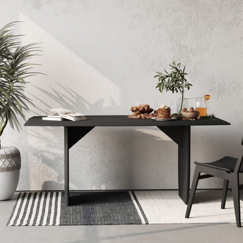 The Adel Dining Table, with its sleek modern black design, is adorned with snacks, a drink, and an open book. It sits beside a black chair on a striped rug, with potted plants in the background.