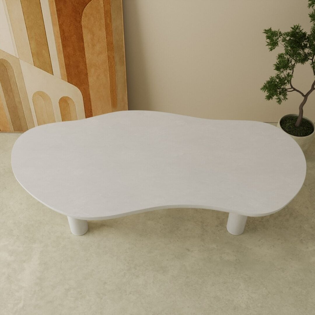 87 Aero Organic Dining Table in white 3 The Aero Organic Dining Table, featuring a white, kidney-shaped top and three cylindrical legs, stands on a light floor beside a plant and abstract wall art.