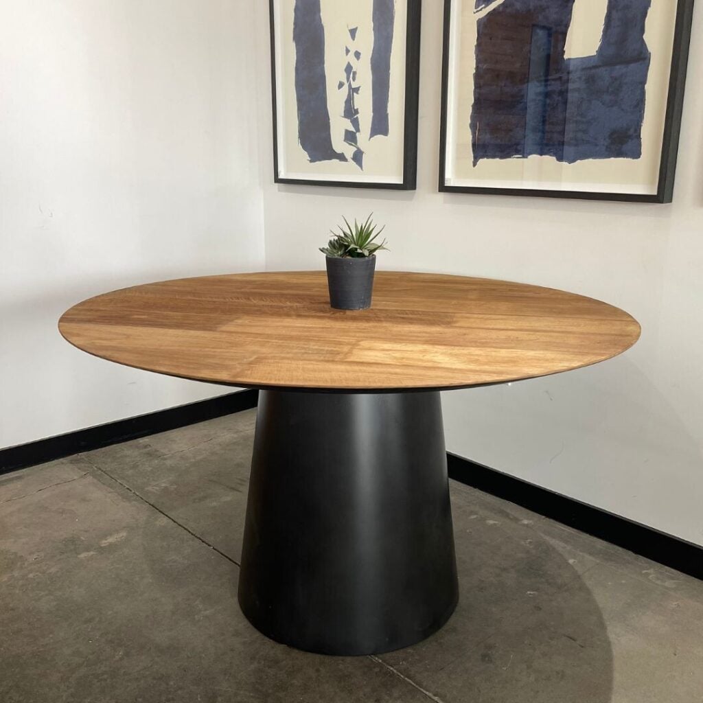 The Atelier Oval Dining Table, featuring a round wooden top and black cylindrical base, is centered in the room with a small potted plant on it. Two pieces of abstract art adorn the wall behind.