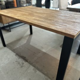 The Metal Dining Table (Natural) features a rectangular wooden top with a textured finish and black metal legs, placed on a concrete floor in a modern indoor space.