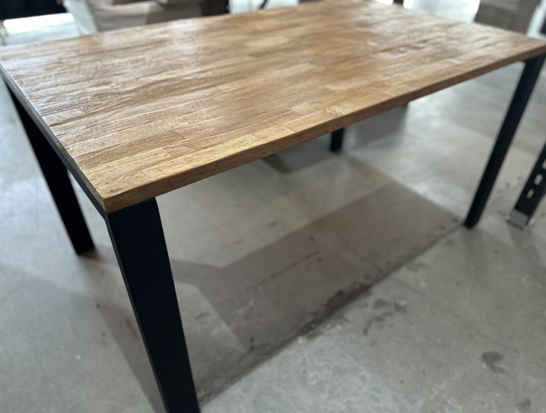 Image_20250806_174109_515 (1) The Metal Dining Table (Natural), featuring a wooden top and black metal legs, stands on a concrete floor in a well-lit room.