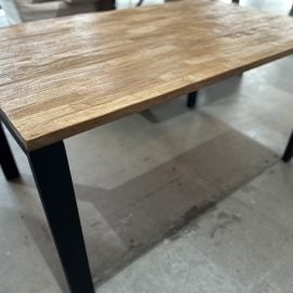 The Metal Dining Table (Natural), featuring a wooden top and black metal legs, stands on a concrete floor in a well-lit room.