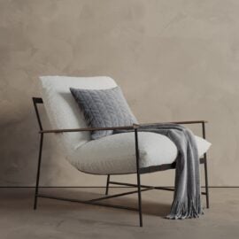 The Aspen 30" Lounge Chair features white upholstery, a black metal frame, a gray quilted pillow, and a gray fringed throw, set against a textured beige wall.