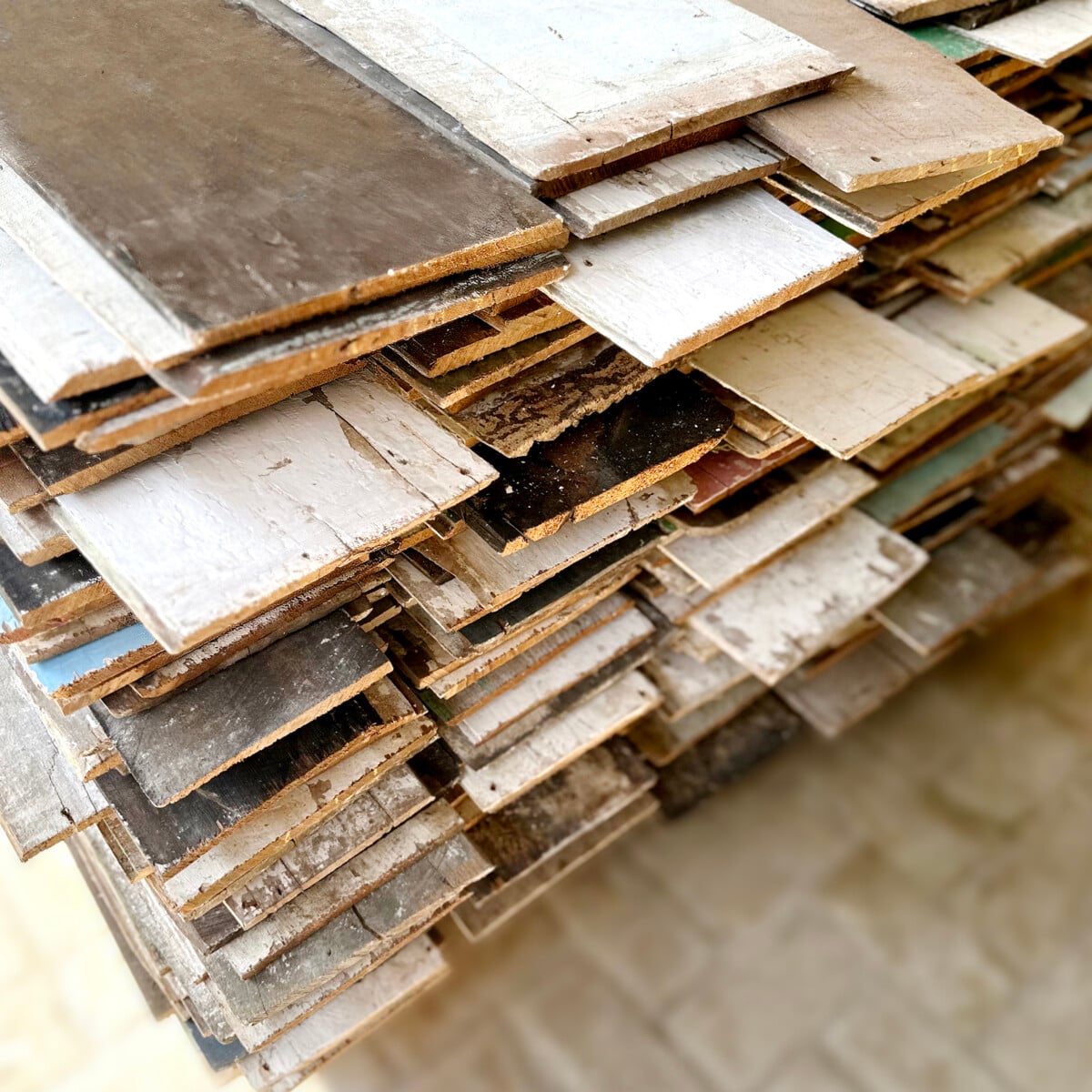 A stack of variously colored and weathered plywood sheets arranged in a pile on a tiled surface.