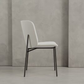 The Kora 20" Dining Chair, featuring modern white upholstery and a black metal frame, sits on a light gray floor against a textured gray wall.