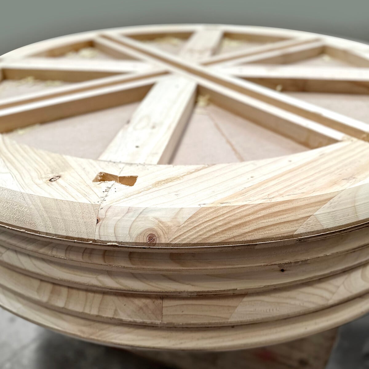 A close-up of a large round wooden tabletop with a geometric pattern and unfinished surface, showing visible wood grain and construction details.