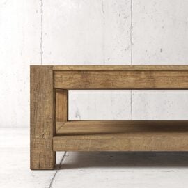 A close-up view of a wooden coffee table with a lower shelf, positioned against a plain, light-colored concrete wall.