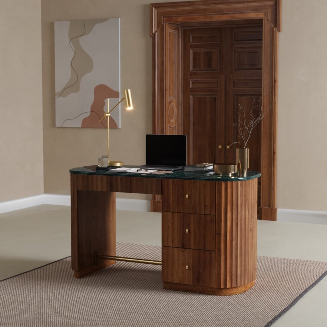 The Elma Desk with drawers holds a laptop, gold lamp, and a vase with a branch. Placed on a rug in front of a wooden door, itโs complemented by abstract art on the wall.