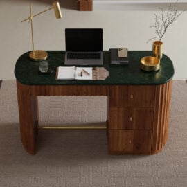 The Elma Desk, featuring a green marble top and three drawers, stands on a beige carpet and holds a laptop, lamp, glass of water, notebook, tray, gold bowl, and a vase with twigs.