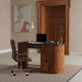 A modern office with the Elma Desk, featuring a green top, paired with a brown office chair, laptop, gold desk lamp, and minimalist decor in a light-walled room with a wooden door.