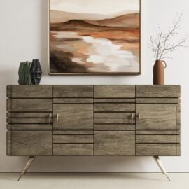 The Kava Sideboard, featuring metal legs, sits against a white wall beneath an abstract landscape painting. Decorative vases and a dried branch are arranged on top.