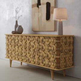 The Kinta Sideboard, featuring a geometric carved design, stands against a light wall and is styled with vases, a decorative branch, lamp, and an abstract painting above.