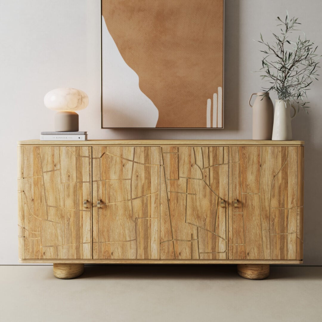 The Noka Sideboard, featuring a textured wood design, stands against a light wall with a lamp, books, and a ceramic vase of greenery on top. Abstract artwork hangs above.