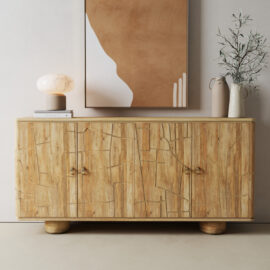 The Noka Sideboard, featuring a textured wood design, stands against a light wall with a lamp, books, and a ceramic vase of greenery on top. Abstract artwork hangs above.
