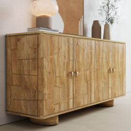The Noka Sideboard, featuring engraved geometric patterns, round handles, and rounded feet, stands against a wall. It is topped with decorative accents like a lamp, books, and vases.