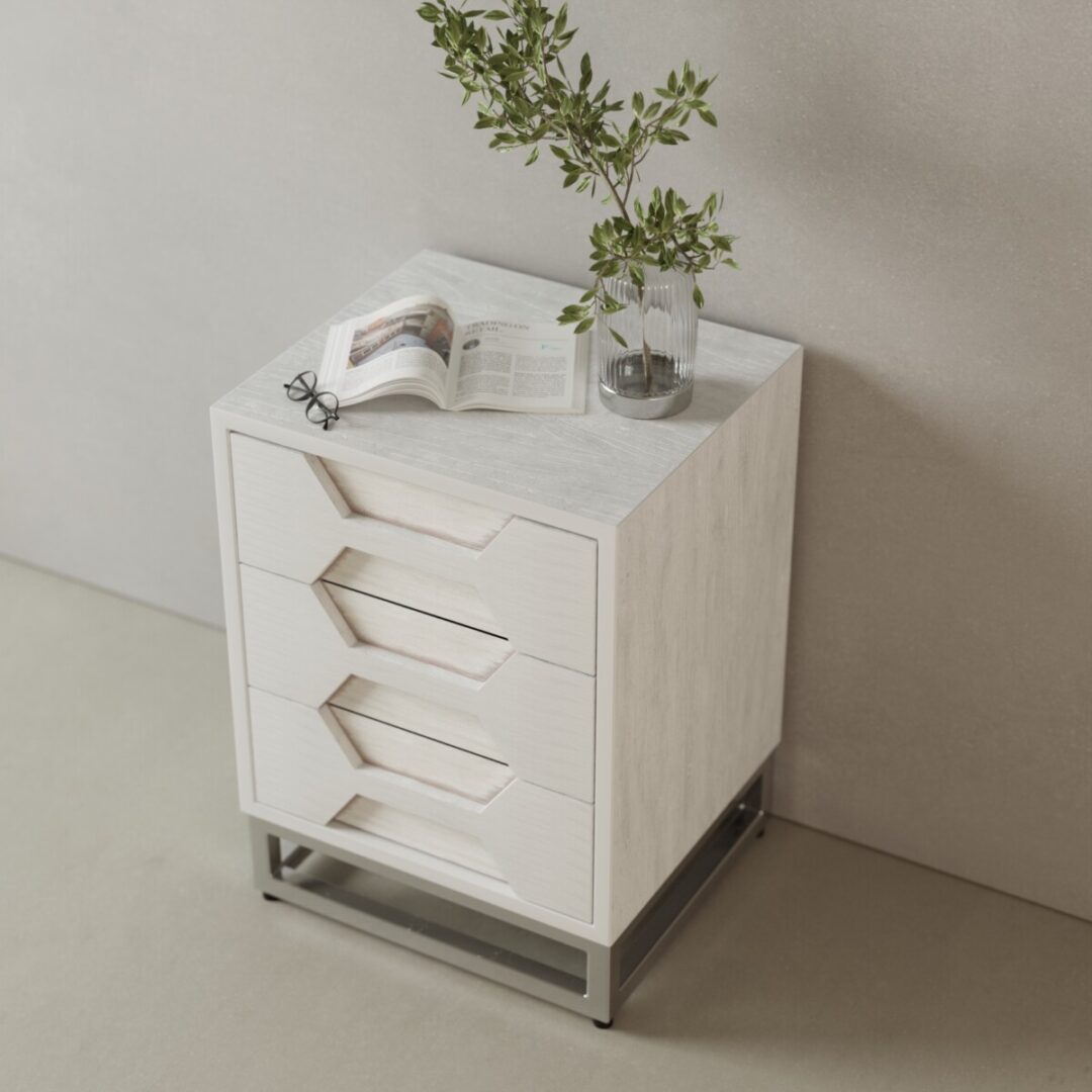 Vexa Nightstand-03 The Vexa Nightstand, a compact white piece with three drawers, is shown against a light wall topped with a glass vase of leafy branches, an open book, and glasses.