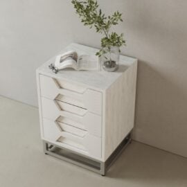 The Vexa Nightstand, a compact white piece with three drawers, is shown against a light wall topped with a glass vase of leafy branches, an open book, and glasses.