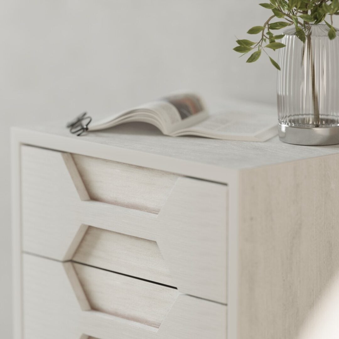 Vexa Nightstand-04 A close-up of the Vexa Nightstand, a modern white wooden drawer unit, with an open magazine and a glass vase with green leaves on top.
