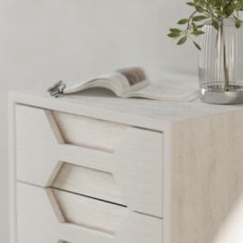A close-up of the Vexa Nightstand, a modern white wooden drawer unit, with an open magazine and a glass vase with green leaves on top.