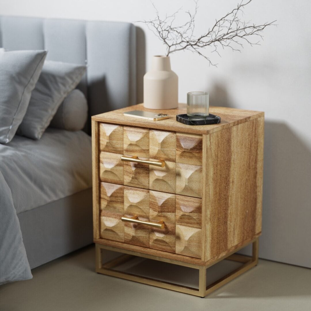 Wadi 18in Nightstand-02 The Wadi Nightstand, featuring geometric-patterned drawers with gold handles and base, sits beside a grey upholstered bed. A beige vase with twigs, a glass, and a tray are arranged on top.
