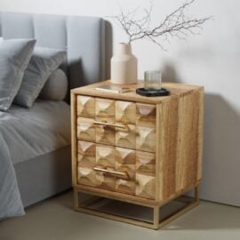 The Wadi Nightstand, featuring geometric-patterned drawers with gold handles and base, sits beside a grey upholstered bed. A beige vase with twigs, a glass, and a tray are arranged on top.