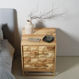 The Wadi Nightstand, featuring a geometric wooden design, holds a beige vase with branches, a smartphone, a tray, and a glass. Part of a bed appears on the left.
