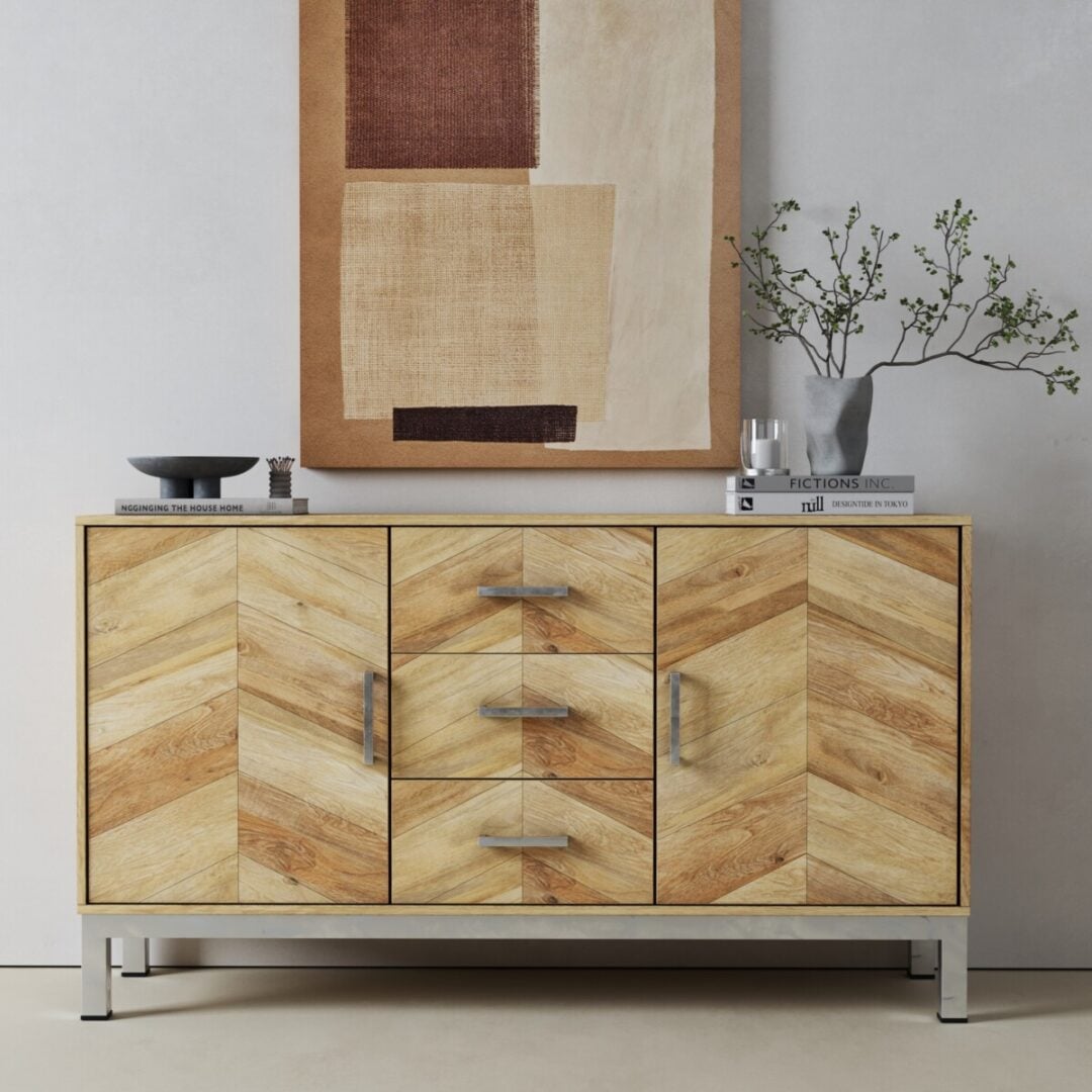 The Zera Sideboard, featuring a herringbone pattern, stands against a white wall topped with books, a bowl, and a vase with branches; an abstract painting hangs above.