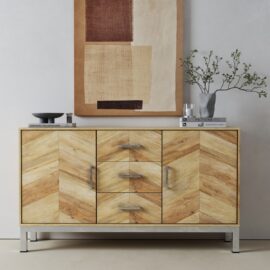 The Zera Sideboard, featuring a herringbone pattern, stands against a white wall topped with books, a bowl, and a vase with branches; an abstract painting hangs above.