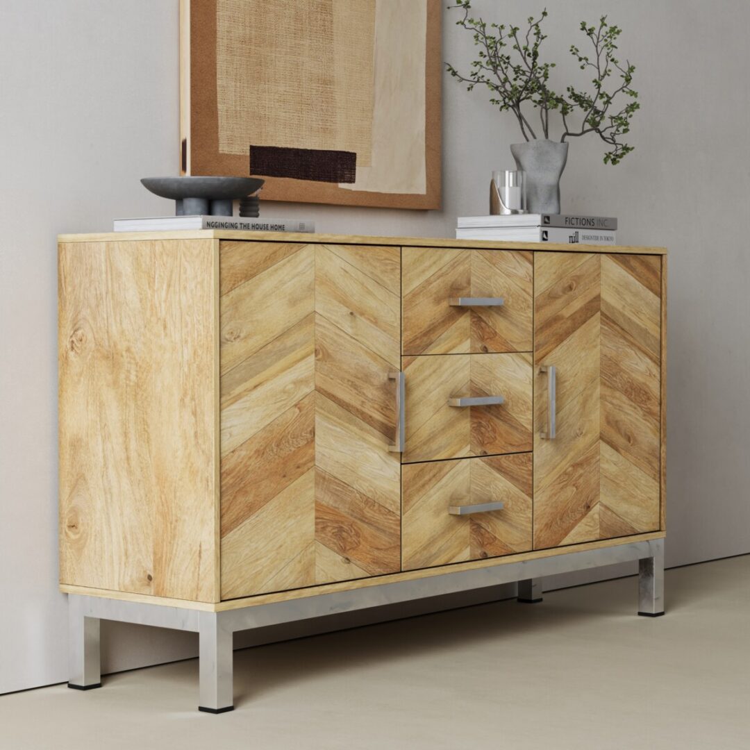 Zera 56in. Sideboard-03 The Zera Sideboard features light wood with a chevron pattern, metal legs, and silver handles. Itโs styled with books, a bowl, a vase of branches on top, and has framed abstract artwork displayed on the wall behind.