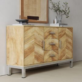 The Zera Sideboard features light wood with a chevron pattern, metal legs, and silver handles. Itโs styled with books, a bowl, a vase of branches on top, and has framed abstract artwork displayed on the wall behind.