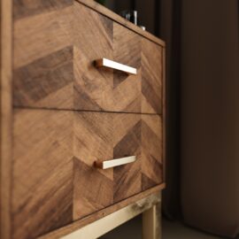 Close-up of the Zera Sideboard, featuring a chevron-patterned wooden finish, two drawers, and gold metal handles, set against a dark background.