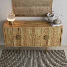 The Luna Sideboard features textured doors and gold handles, styled with a lamp, vase, and decor on top. A woven wall hanging above and a patterned rug below complete the look.