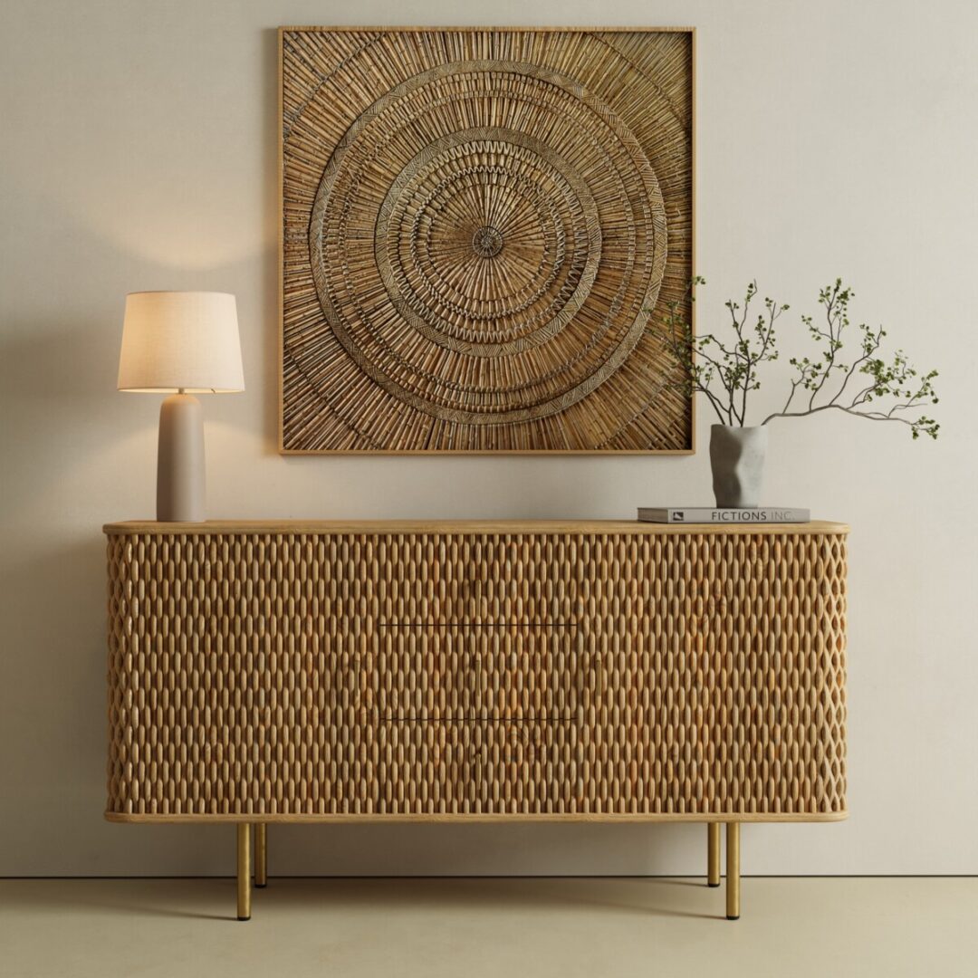 A wicker sideboard with a lamp, a vase with branches, and books sits below a large woven wall art piece in a minimalist, neutral-toned room.