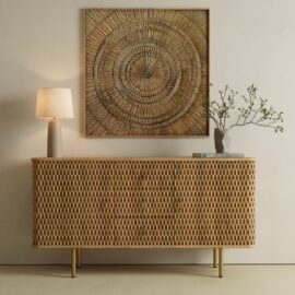 A wicker sideboard with a lamp, a vase with branches, and books sits below a large woven wall art piece in a minimalist, neutral-toned room.