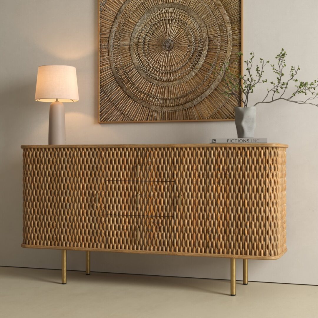 Salaro 60in Sideboard-02 A woven rattan sideboard with gold legs stands against a beige wall, topped with a lamp, a vase with branches, and books; a large circular wicker wall art hangs above.