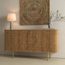 A woven rattan sideboard with gold legs stands against a beige wall, topped with a lamp, a vase with branches, and books; a large circular wicker wall art hangs above.