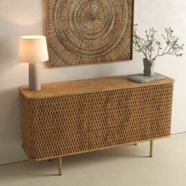 A wooden sideboard with a textured front, a lamp, a gray vase with branches, and a book placed on top, beneath a circular woven wall art.